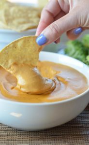 A vertical shot of pretty lady fingers with blue nails dipping a tortilla chip into a bowl of cashew queso.