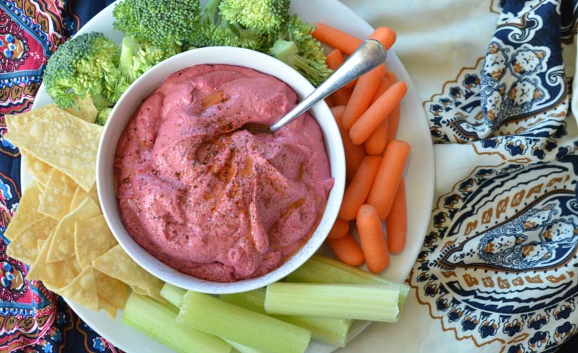 Bowl of bright red beet hummus surrounded by vegetables and tortilla chips.